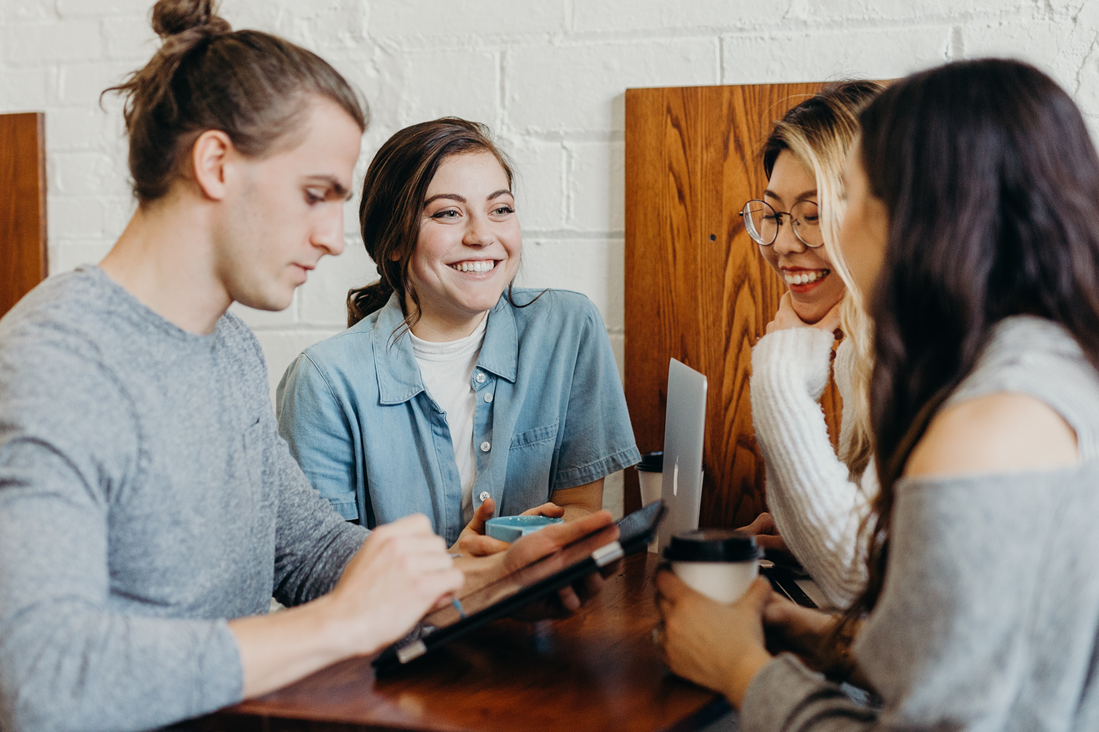 Four people leaning on a wooden table talking, drinking hot drinks and using tablets.