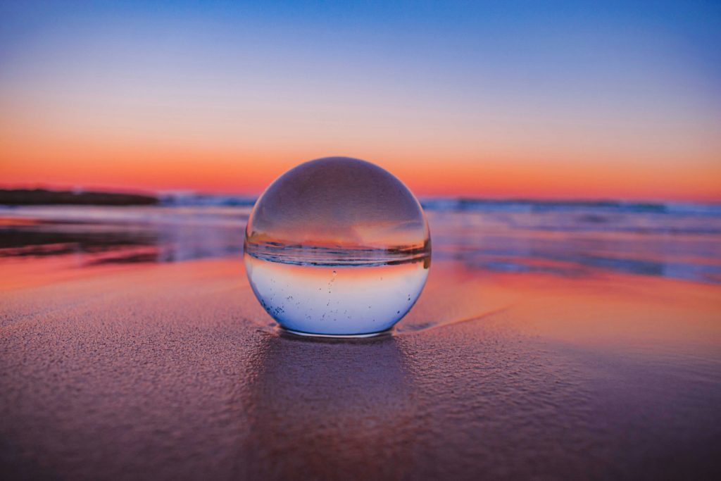 A clear glass sphere rests on wet sand at the beach during sunset. The sphere reflects the vivid colours of the sky, creating an inverted image of the horizon. The background shows calm ocean waves and a gradient sky transitioning from deep blue at the top to warm orange and pink near the horizon.